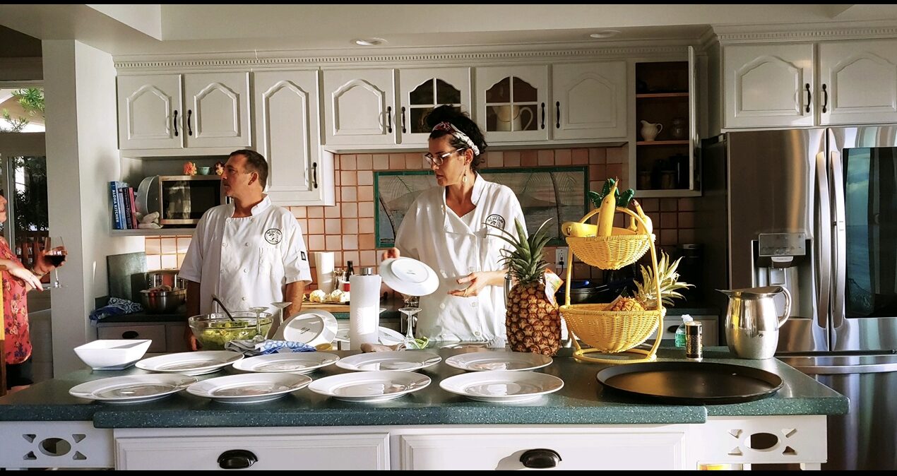 Val and John in chef whites, plating dinner for a group in a guest's kitchen
